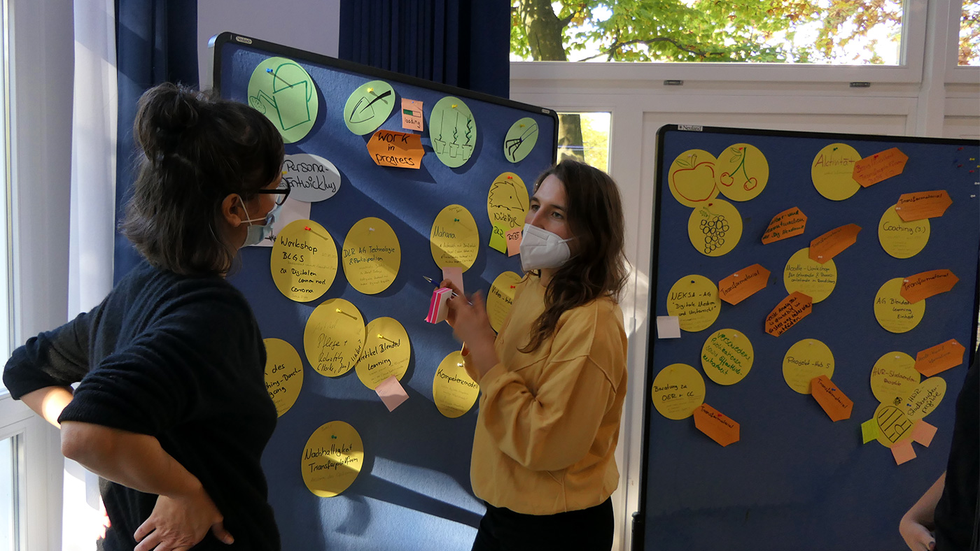 2 Personen stehen vor einer Metaplanwand unt sprechen miteinander - Schulungssituation Digitale Akademie Pflege 4.0 Projekttreffen Oktober 2021